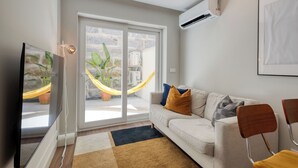 A modern living room with a sofa, a TV, a hammock, and a sliding glass door leading to a balcony with plants.