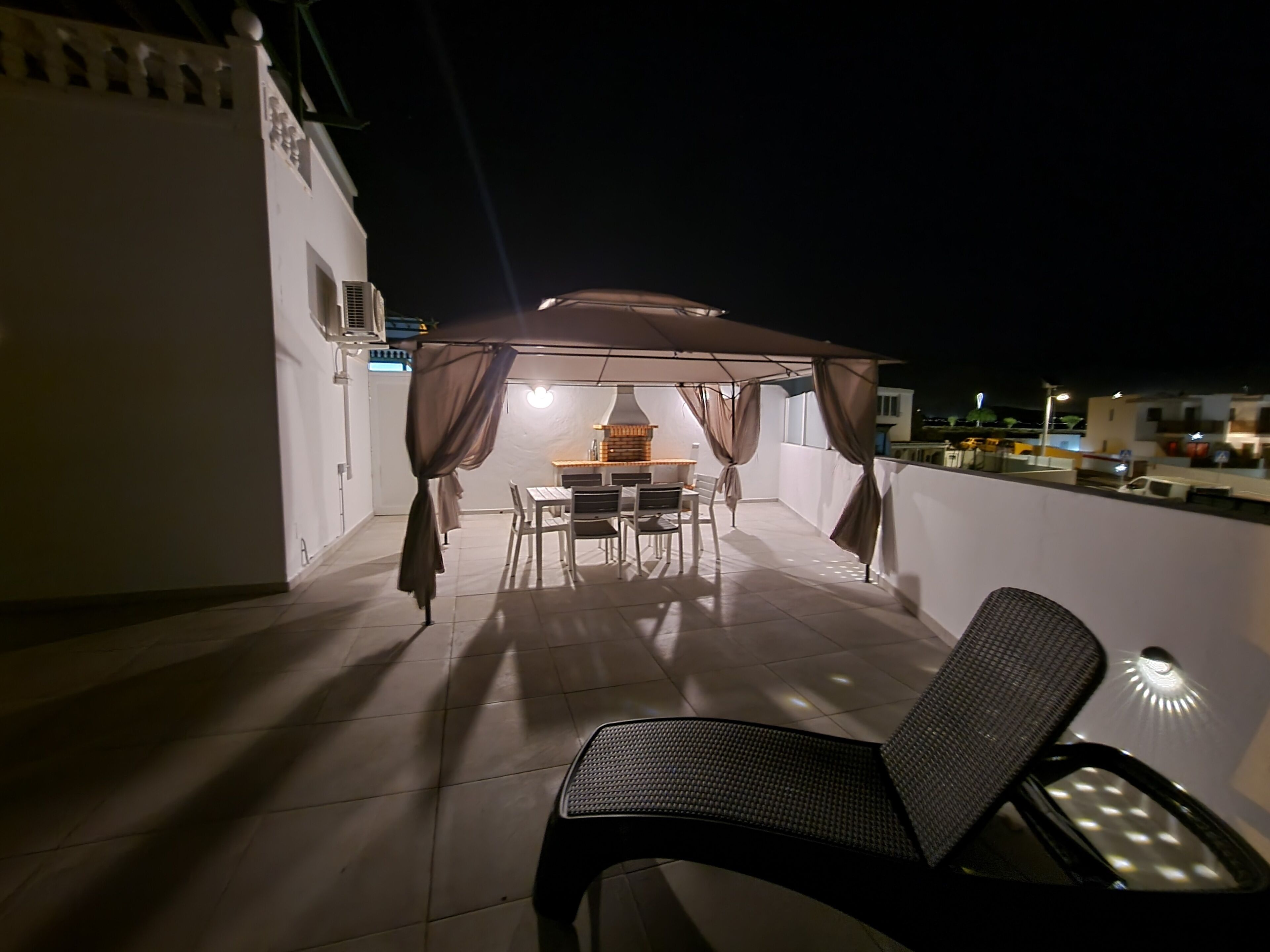 Een overdekt terras met een tafel, stoelen en een comfortabele fauteuil, plus een wandlamp.