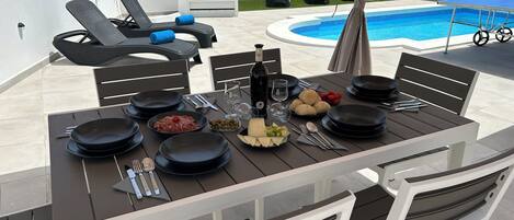 Mesa de comedor lista para una comida, con platos, cubiertos y una botella de vino, con vista a una piscina y sillas de descanso.