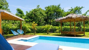 Une piscine dotée d’une terrasse en bois et de chaises longues, entourée d’une végétation luxuriante et agrémentée d’un gazebo à toit de chaume.