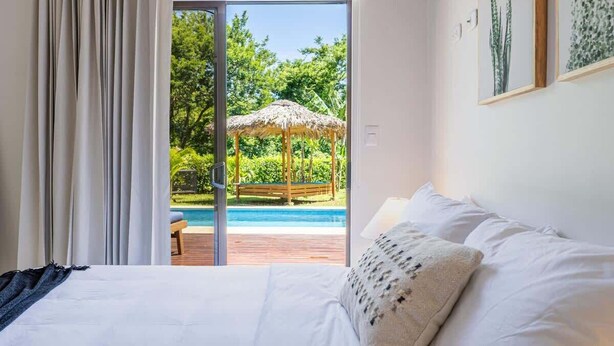 Une chambre avec un grand lit, une vue sur une piscine et un gazebo à toit de chaume à l’extérieur.