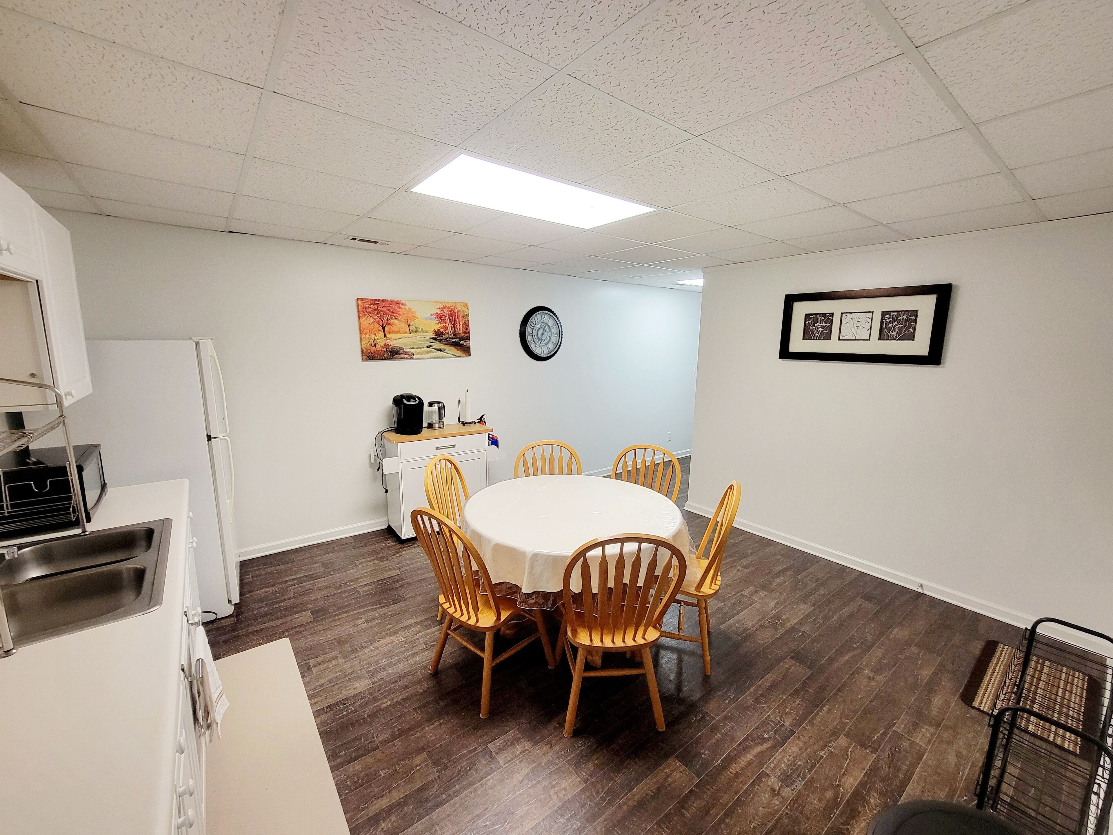 A dining area with a round table and wooden chairs, a kitchen area with a refrigerator and microwave, and a clock on the wall.