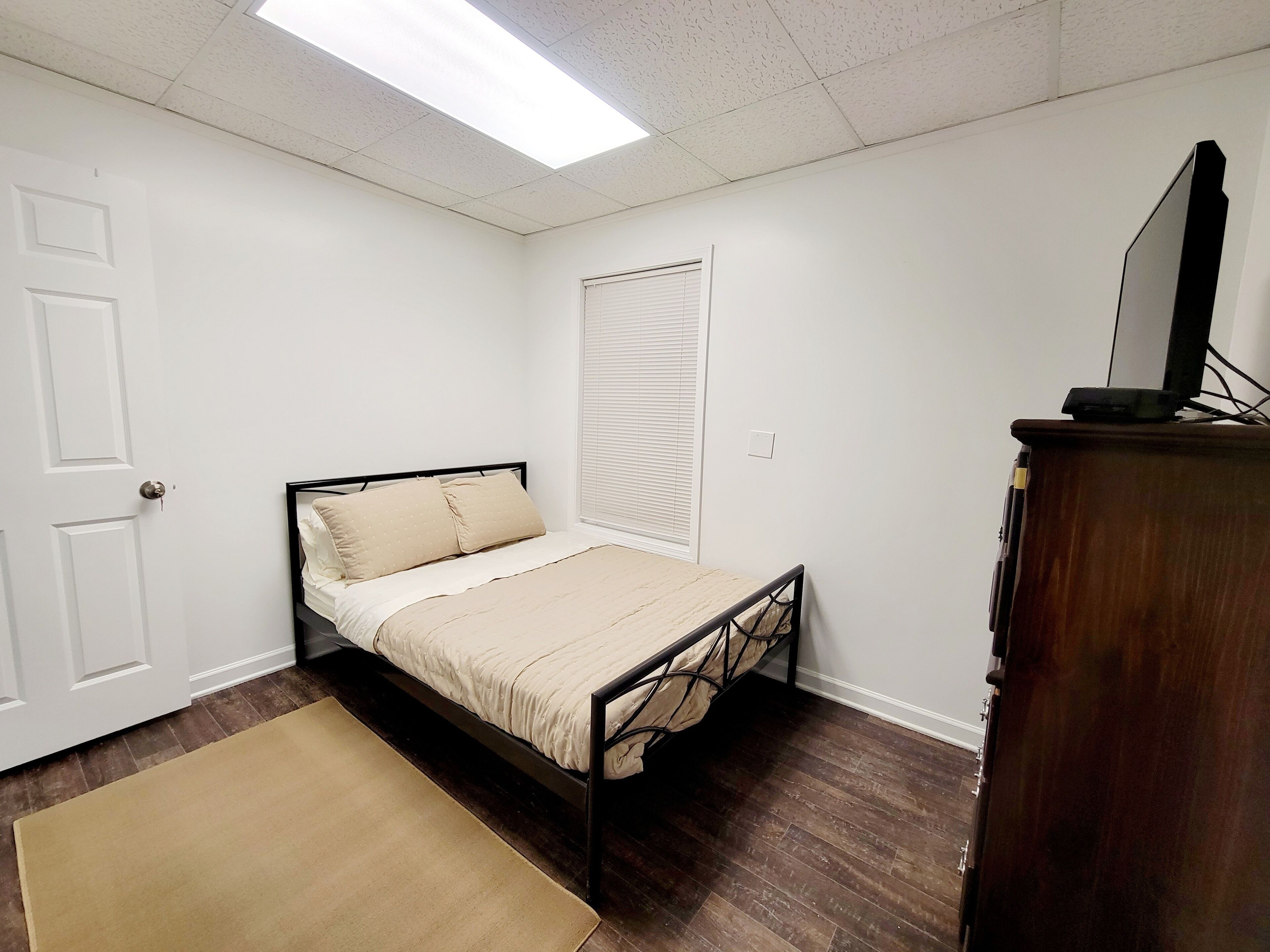 Un dormitorio con cama, cómoda de madera, televisor encima y ventana con persianas.