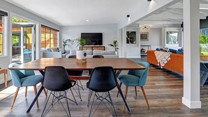 Zona pranzo moderna con tavolo in legno e sedie nere, un divano grigio e una TV sullo sfondo.