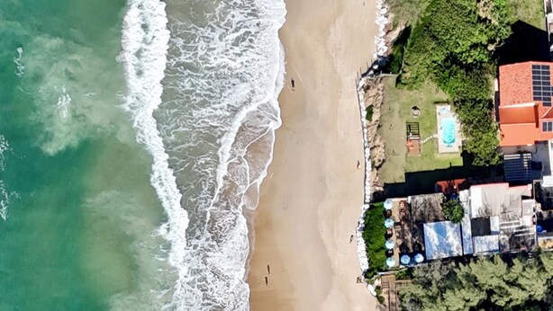 An aerial view of a beach with waves, a few people, and a residential area with houses and greenery. - Beachfront house with spa! Fire pit / Spa / Hammocks / Gardens / Parking (Morro das Pedras Florianópolis)