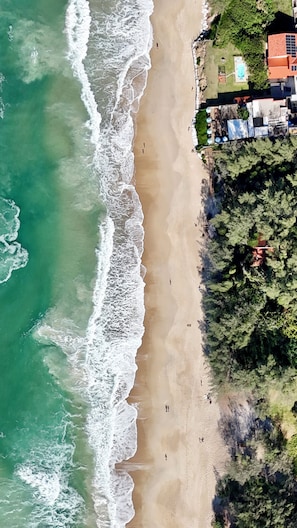 Une vue aérienne d’une plage avec des vagues, quelques personnes et une zone résidentielle avec des maisons et de la végétation.