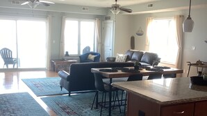 Un vaste espace de vie comprenant un îlot de cuisine, une table à manger et des chaises. On y trouve également des ventilateurs de plafond, de grandes fenêtres avec des rideaux et une vue sur l’extérieur.