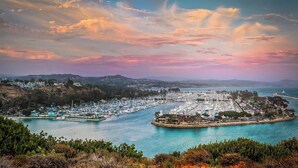 A coastal marina with boats docked, a small island with buildings, and a hilly landscape in the background.