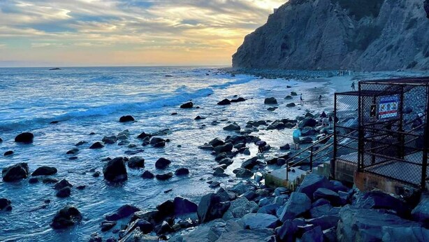 A rocky beach with a steep cliff, a fenced area, and a clear sky.