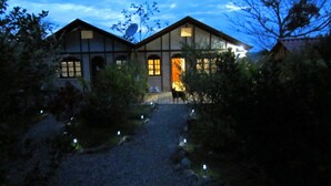 Une maison traditionnelle de style japonais, avec un chemin de gravier menant à l’entrée, entourée d’arbres et illuminée de petites lumières.