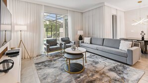 A modern living room with a grey sofa, a round coffee table, and a large window with curtains. - Luxury 4BR Townhome w/ Pool Near Disney (Kissimmee)