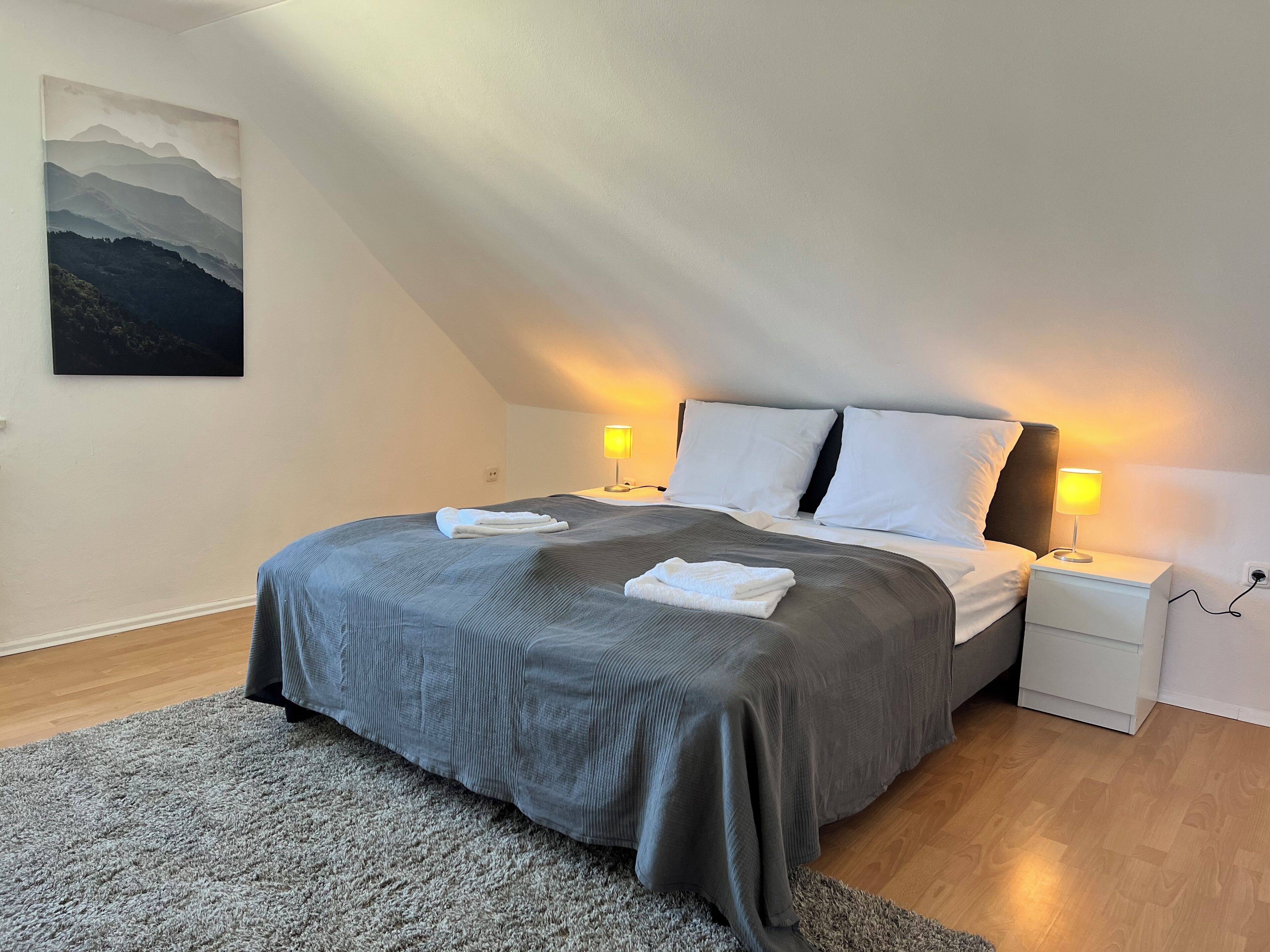 Una cama bien hecha con cubrecama gris y almohadas blancas, dos lámparas de noche, una mesita de noche pequeña y una alfombra gris sobre piso de madera.