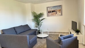 A modern living room with a grey sofa, a grey armchair, a small round table, a potted plant, and a flat-screen TV on a stand. - ~straubings Großes Apartment~ (Straßkirchen)