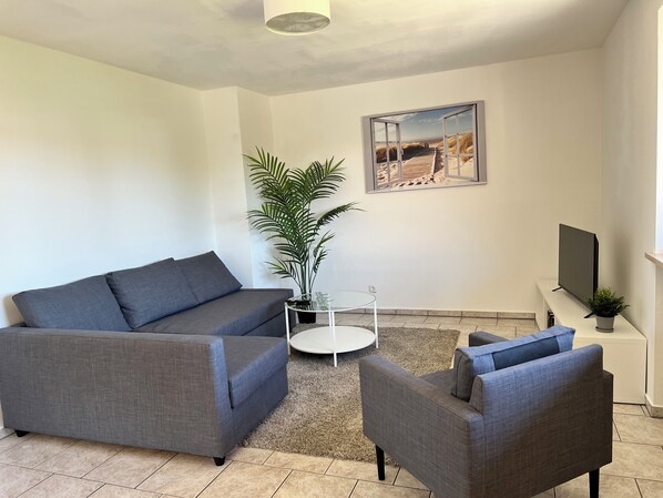 A modern living room with a grey sofa, a grey armchair, a small round table, a potted plant, and a flat-screen TV on a stand. - ~straubings Großes Apartment~ (Straßkirchen)