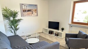 A modern living room with a grey sofa, a small round coffee table, a flat-screen TV on a white stand, and a large window with a view of the outdoors. - ~straubings Großes Apartment~ (Straßkirchen)
