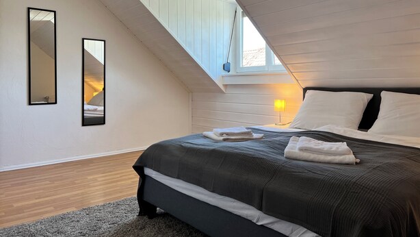 A bedroom with a sloped ceiling, a bed with dark bedding, a bedside lamp, and two mirrors on the wall. - ~straubings Großes Apartment~ (Straßkirchen)