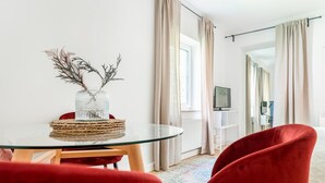 Ein modernes Wohnzimmer mit Glastisch, roten Stühlen, einer Glasvase mit Ästen und einem Fernseher auf einem Schrank.