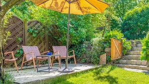 Eine Terrasse mit zwei Korbstühlen und einem Tisch, einem gelben Sonnenschirm und einer Steinmauer mit einem Holztor.