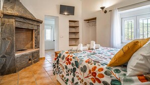 A bedroom with a stone fireplace, a bed with a floral bedspread, a TV mounted on the wall, and a window with curtains. - Stunning home in Torrox (Torrox)