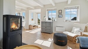 A spacious living room with a fireplace, wooden flooring, a sofa, a dining table, and a view of the outdoors. - 2 bedroom stunning home in Blåvand (Blåvand)