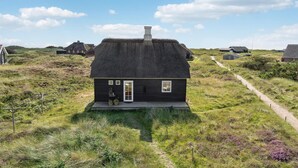 A thatched-roof house with a wooden exterior, surrounded by grassy fields and other similar houses. - 2 bedroom stunning home in Blåvand (Blåvand)