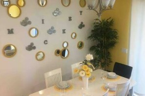 Comedor con una mesa dispuesta para cuatro personas, rodeada de sillas. En la pared, espejos circulares y letras que deletrean un nombre.