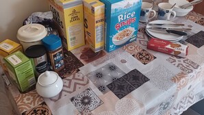 Ein Esstisch mit verschiedenen Speisen und Küchenutensilien, darunter Schachteln mit Frühstücksflocken, eine Packung Reissnacks und ein Honigglas.