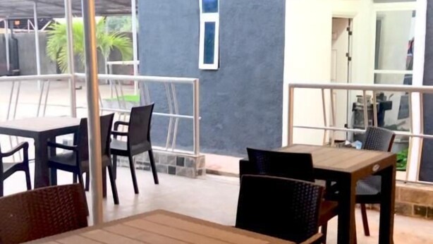 Un área de estar al aire libre con mesas y sillas, una sección cubierta con techo metálico y vista a un edificio con fachada oscura.