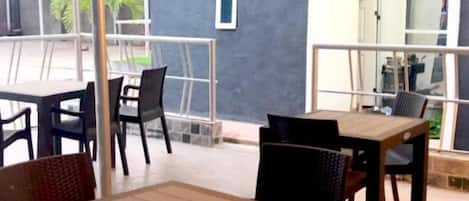 Un área de estar al aire libre con mesas y sillas, una sección cubierta con techo metálico y vista a un edificio con fachada oscura.