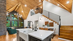 A spacious kitchen with a large island, a dining area with wicker furniture, and a staircase leading to an upper level. - Lazy Day Hideaway Cabin | Modern-Boho Retreat (Broken Bow)