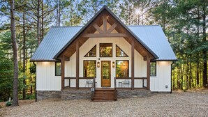 A cabin with a stone foundation, wooden beams, and a metal roof, surrounded by trees. - Lazy Day Hideaway Cabin | Modern-Boho Retreat (Broken Bow)