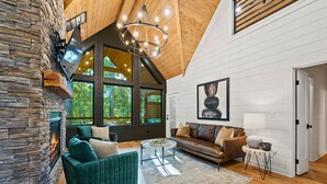 A spacious living room with a stone fireplace, a large window, and a cozy seating area. - Lazy Day Hideaway Cabin | Modern-Boho Retreat (Broken Bow)