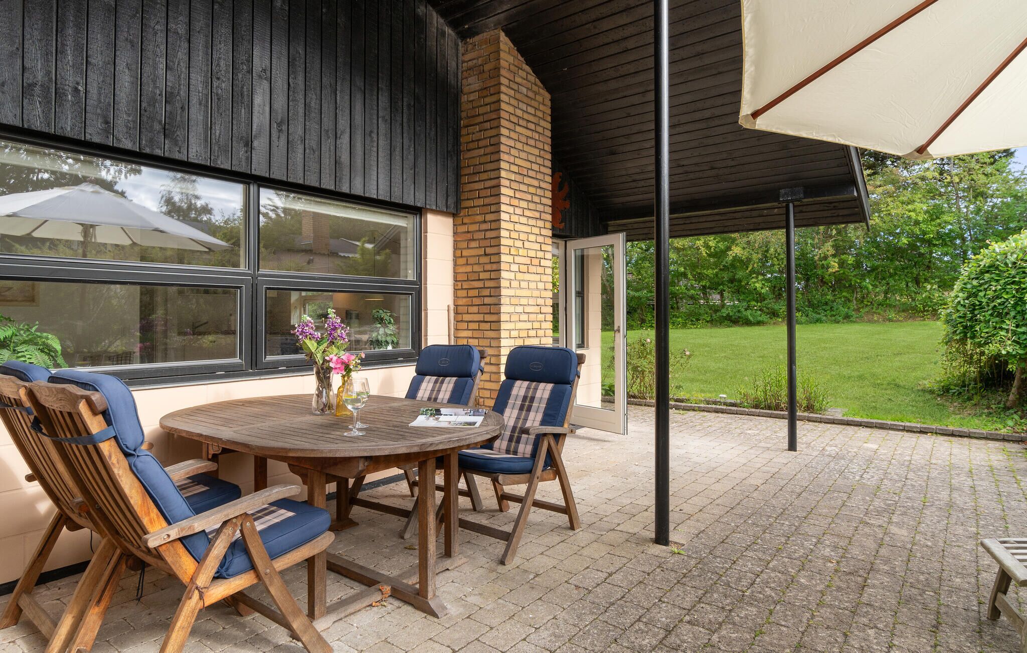 Een terras buiten met een ronde houten tafel en stoelen, een bakstenen schoorsteen en uitzicht op een groene tuin.