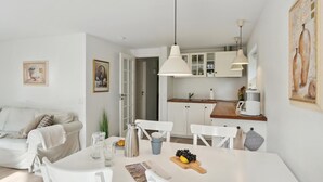 A modern, well-lit living room with a white sofa, a dining table with a fruit centerpiece, and a kitchen area with white cabinets and a coffee machine.