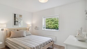 A bedroom with a bed, bedside lamps, a window with a view of greenery, and a small white table with a vase.