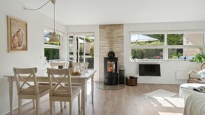 A cozy living room with a fireplace, a dining area with a table and chairs, and large windows offering a view of the garden.