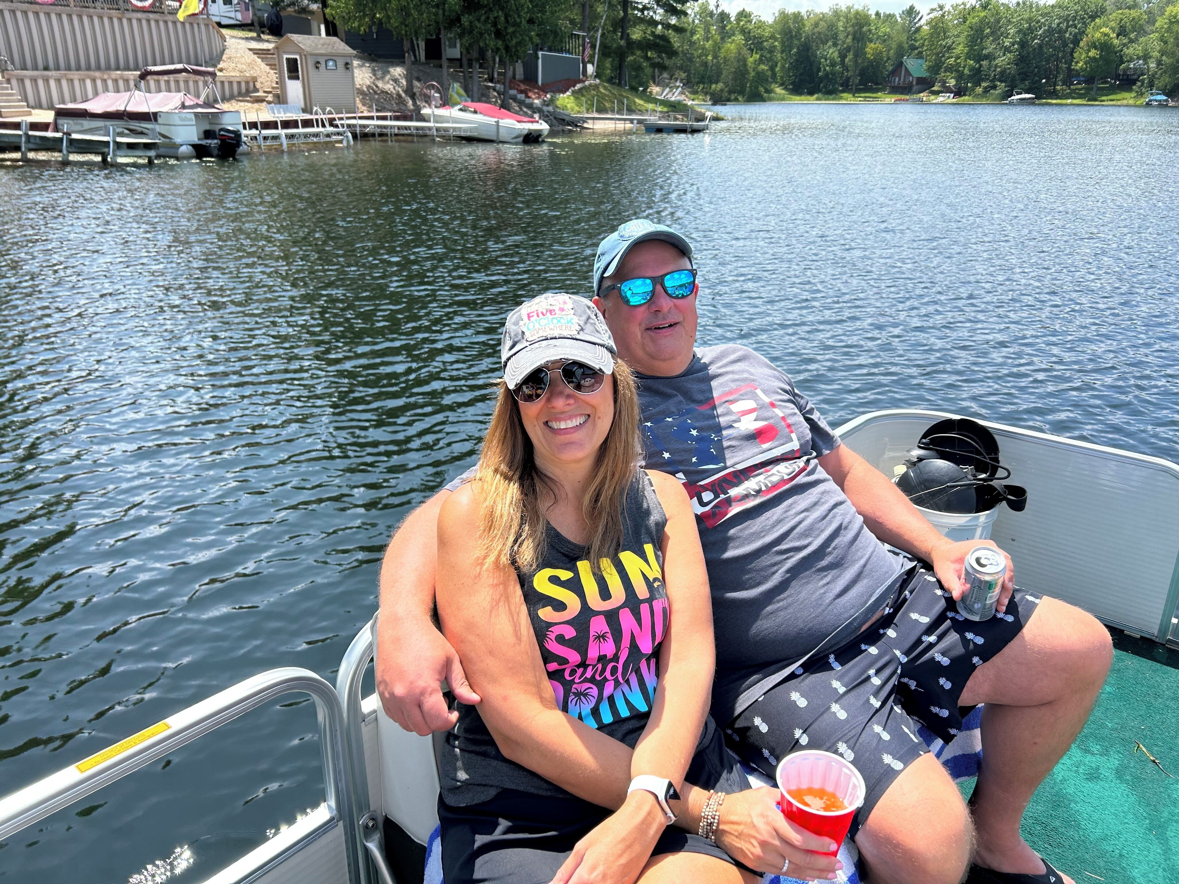 A couple on a boat with a view of a calm lake and docked boats in the background.