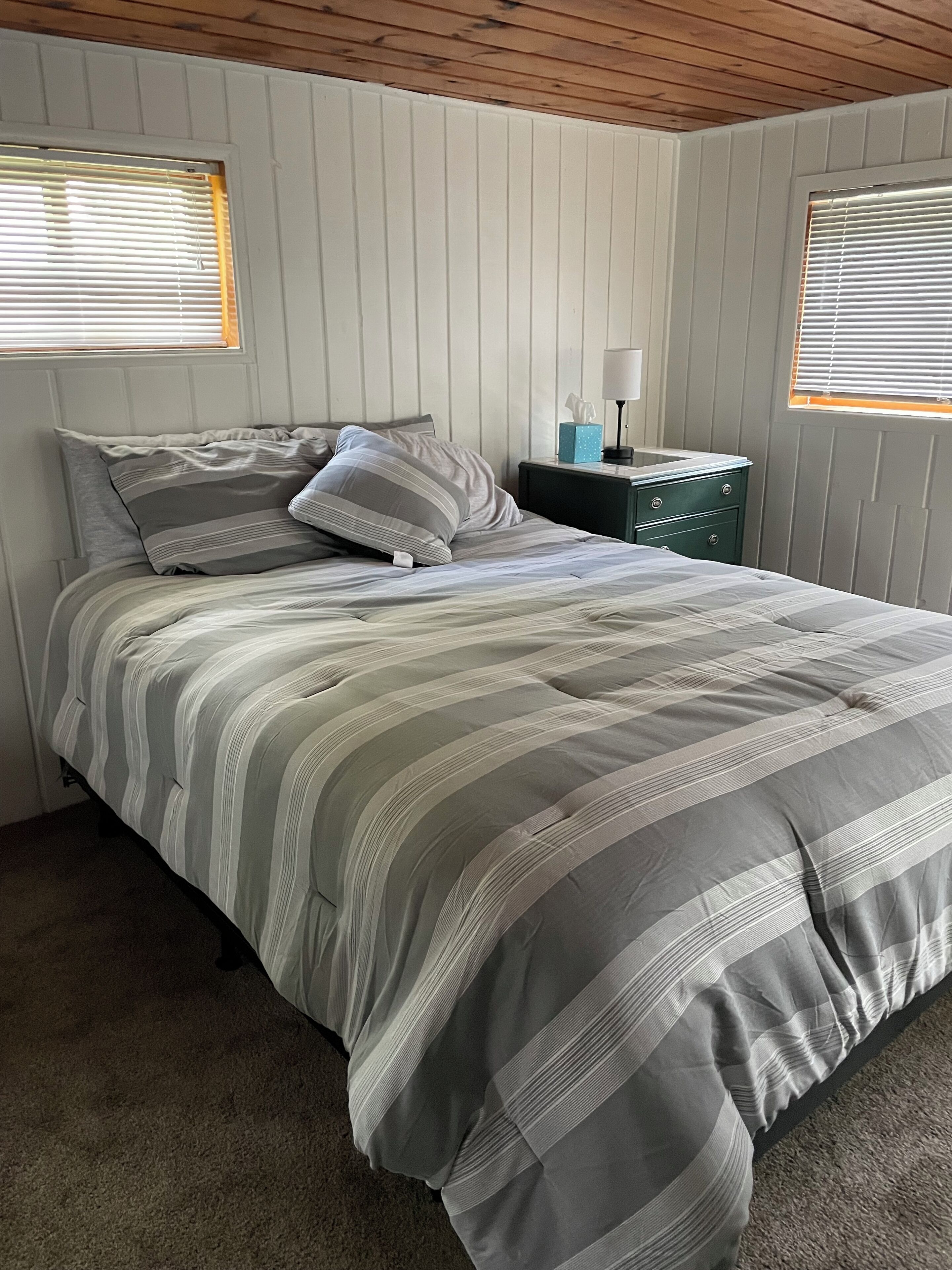 Una cama bien hecha con ropa de cama a rayas, una mesita de noche con lámpara y una ventana con persianas.