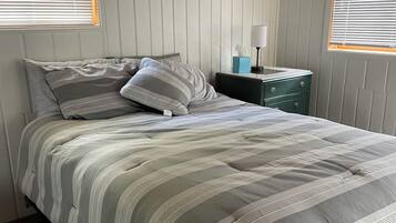 Una cama bien hecha con ropa de cama a rayas, una mesita de noche con lámpara y una ventana con persianas.
