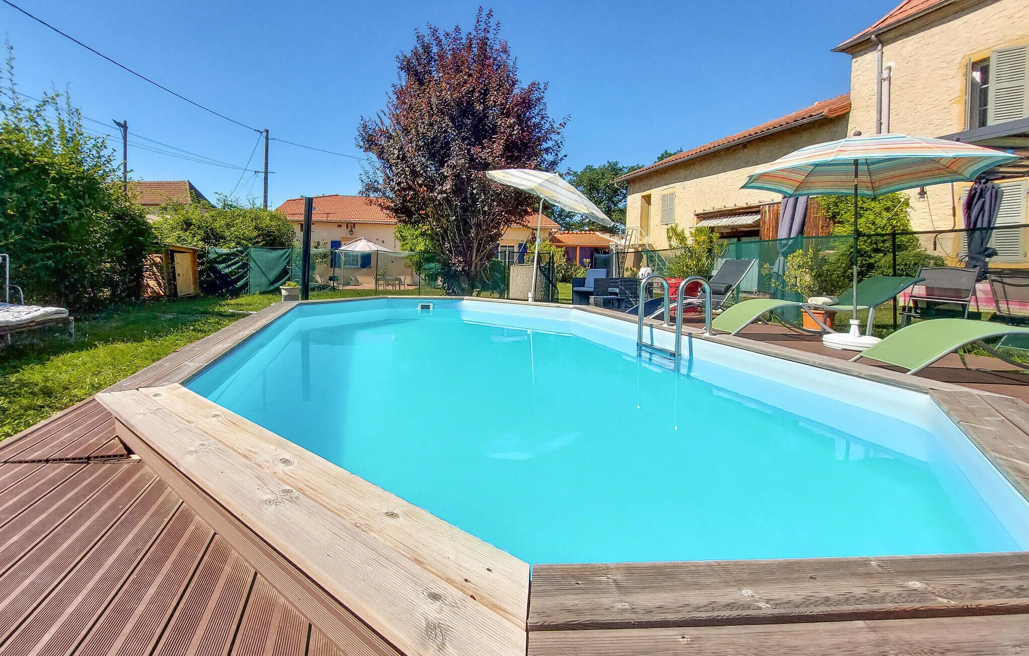 A rectangular swimming pool with a wooden deck, surrounded by a garden and a house with a tiled roof.