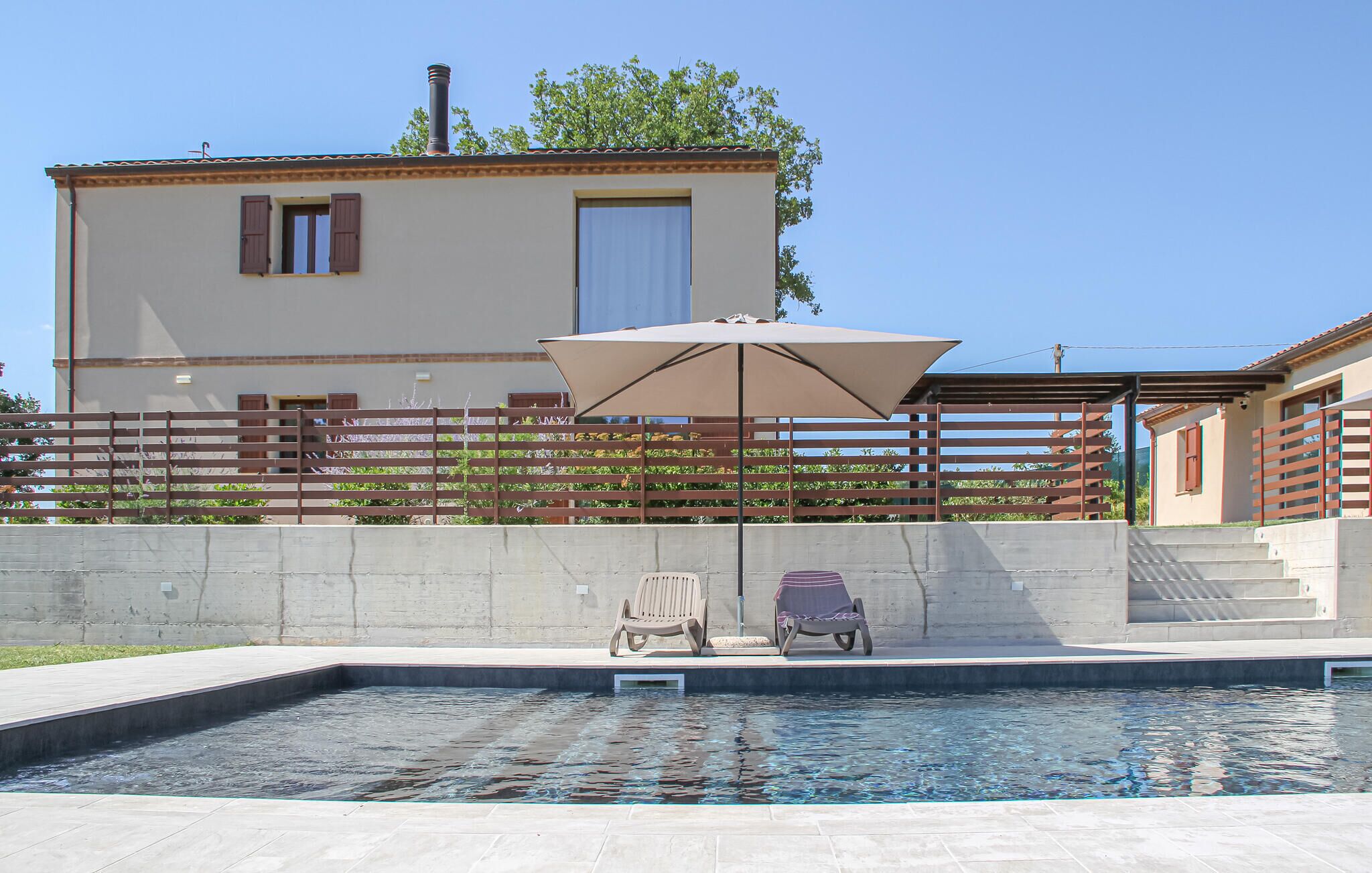 Una piscina con dos sillas de descanso, un pérgola con toldo y un edificio con tejado de tejas al fondo.