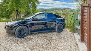 Un coche eléctrico elegante y de color oscuro estacionado en un camino de grava, con una valla verde y árboles al fondo.