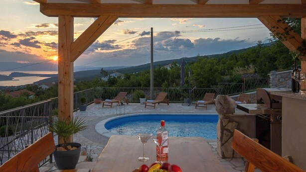 Eine Holzterrasse mit Pool, Liegestühlen, einem Tisch mit Früchten und Getränken und einem malerischen Ausblick auf Hügel und einen Sonnenuntergang.