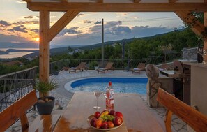 A wooden deck with a pool, lounge chairs, a table with fruit and drinks, and a scenic view of hills and a sunset. - Cozy home in Jadranovo with sauna (Jadranovo)