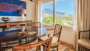 Un comedor con una mesa de vidrio, sillas de madera, un cuadro de un pez, un cuenco de fruta y vistas a un paisaje montañoso.