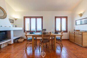 A dining area with a wooden table set for four, surrounded by wicker chairs. There are two windows with wooden frames, a woven wall hanging, and a wicker cabinet to the right.