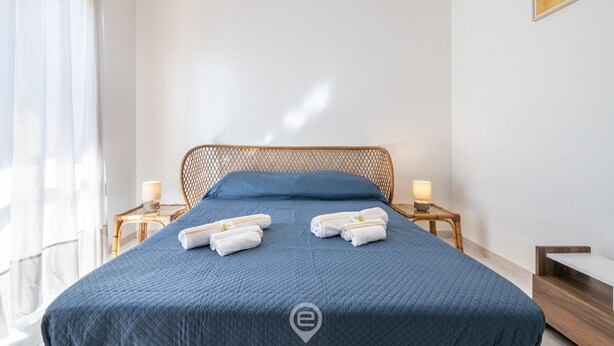 A neatly made bed with blue bedding and two rolled towels, a wooden headboard, bedside tables with lamps, and a window with sheer curtains. - Sunset Oasis Apartment (Quartu Sant'Elena)