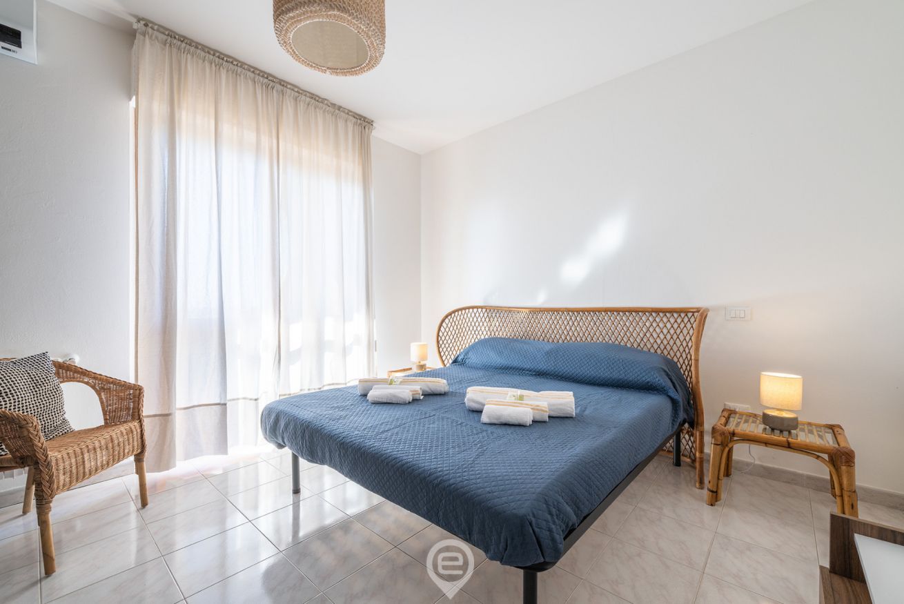 Un dormitorio con cama, una silla, una mesita de noche y un ventanal con cortinas.