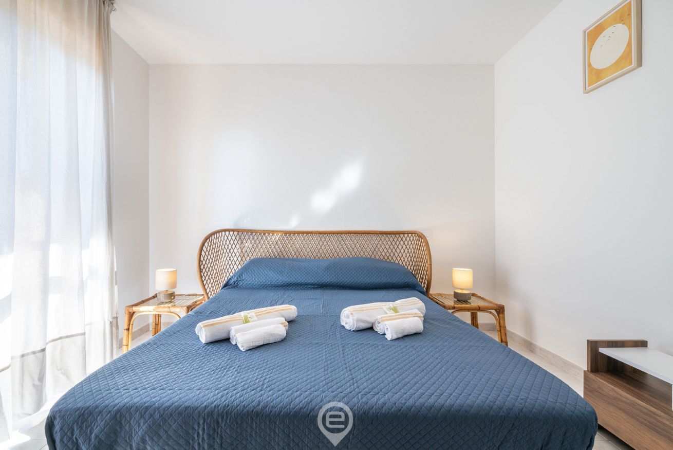 Un lit bien fait, avec une literie bleue et deux serviettes enroulées, une tête de lit en bois, des tables de chevet avec des lampes et une fenêtre ornée de rideaux légers.
