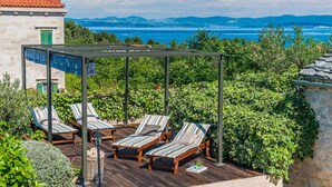 Een overdekt terras met houten ligbedden en een pergola, met uitzicht op een prachtig zee- en berglandschap.